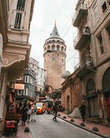 A bustling street scene with a historical tower rising prominently in the background. The streets are narrow and lined with old buildings, featuring balconies and intricate architectural details. Various people are walking, interacting, and exploring the area. On the side, there is a cozy caf&eacute; adorned with lights, adding a touch of warmth to the scene. A yellow taxi and a motorcycle can be seen navigating the inclined road.
