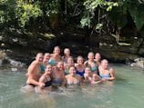 A group of women are standing together in clear water, smiling broadly. They are surrounded by lush greenery and rocks, suggesting a natural setting. Sunlight filters through the leaves, creating a warm and inviting atmosphere.