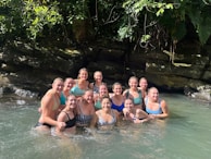 A group of women are standing together in clear water, smiling broadly. They are surrounded by lush greenery and rocks, suggesting a natural setting. Sunlight filters through the leaves, creating a warm and inviting atmosphere.