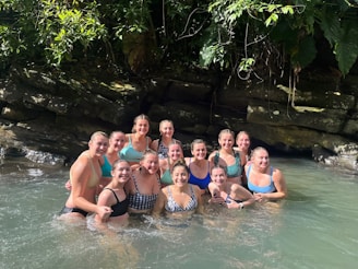 A group of women are standing together in clear water, smiling broadly. They are surrounded by lush greenery and rocks, suggesting a natural setting. Sunlight filters through the leaves, creating a warm and inviting atmosphere.
