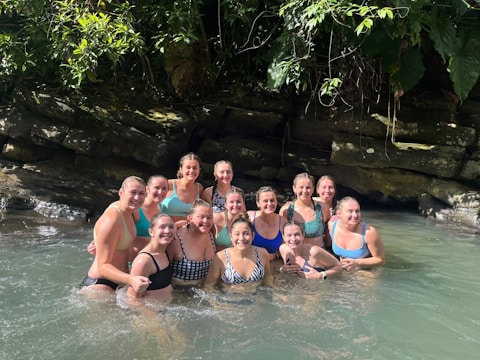 A group of women are standing together in clear water, smiling broadly. They are surrounded by lush greenery and rocks, suggesting a natural setting. Sunlight filters through the leaves, creating a warm and inviting atmosphere.