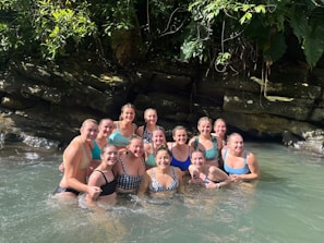 A group of women are standing together in clear water, smiling broadly. They are surrounded by lush greenery and rocks, suggesting a natural setting. Sunlight filters through the leaves, creating a warm and inviting atmosphere.