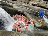 A group of people in swimsuits are gathered in a rocky area underneath a waterfall, surrounded by rushing water and large rocks. One person is standing on a rock beside the group, overlooking the scene, while everyone else is either standing or sitting in the shallow water.