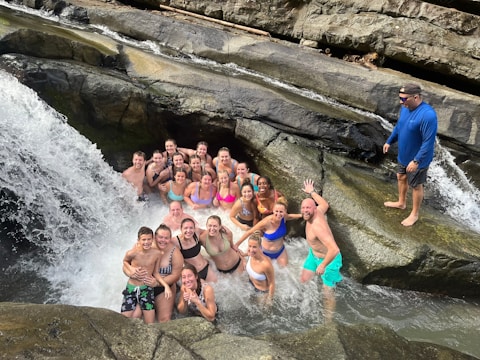 A group of people in swimsuits are gathered in a rocky area underneath a waterfall, surrounded by rushing water and large rocks. One person is standing on a rock beside the group, overlooking the scene, while everyone else is either standing or sitting in the shallow water.
