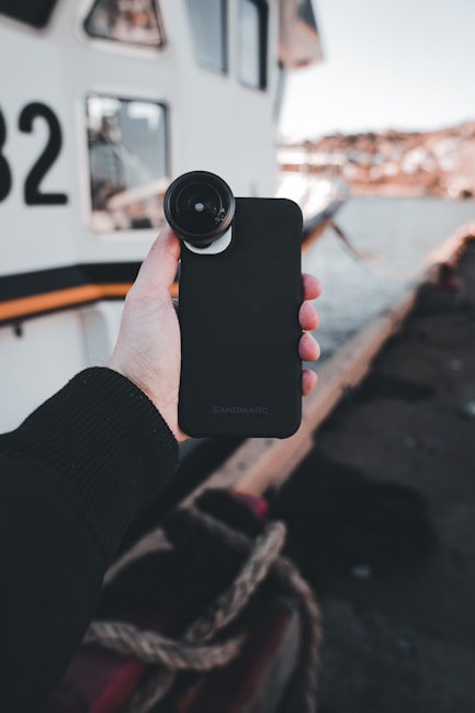 A hand is holding a smartphone with a camera lens attachment in front of a boat docked by a waterfront. The scene includes a section of the boat with the number 82 visible, and there are ropes and part of the dock in the foreground.