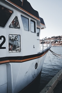 A close-up view of a docked boat with a white hull featuring black and orange accents. The boat displays several windows and has a partially visible number '2'. The background includes a calm body of water and a distant shoreline with some buildings.