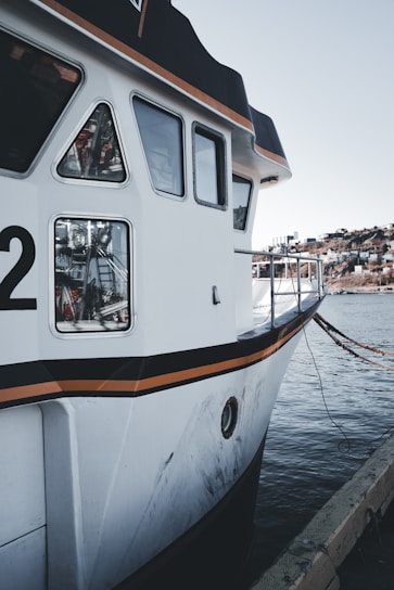 A close-up view of a docked boat with a white hull featuring black and orange accents. The boat displays several windows and has a partially visible number '2'. The background includes a calm body of water and a distant shoreline with some buildings.