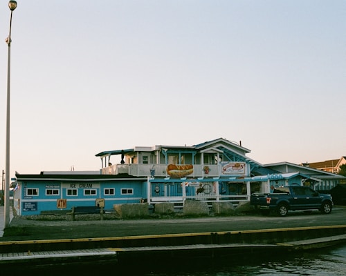 A blue waterfront building resembling a restaurant or cafe, decorated with signs for ice cream and a patio bar. A pickup truck is parked nearby, and the area seems quiet and serene with a clear sky overhead.