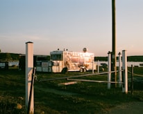 A shiny food trailer is parked on grassy terrain near a body of water during sunset. The trailer reflects the warm light, and there are signs and white posts surrounding the area. A wooden pole stands nearby, and distant trees line the horizon.