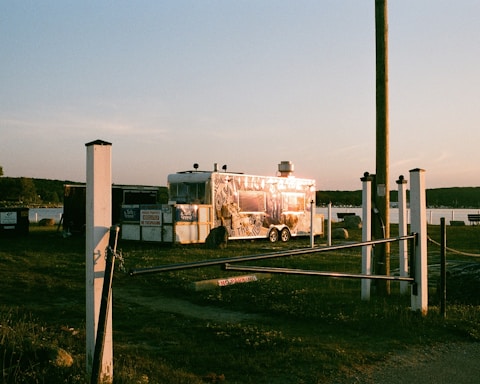 A shiny food trailer is parked on grassy terrain near a body of water during sunset. The trailer reflects the warm light, and there are signs and white posts surrounding the area. A wooden pole stands nearby, and distant trees line the horizon.
