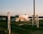A shiny food trailer is parked on grassy terrain near a body of water during sunset. The trailer reflects the warm light, and there are signs and white posts surrounding the area. A wooden pole stands nearby, and distant trees line the horizon.