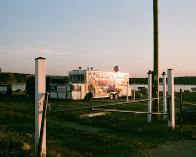 A shiny food trailer is parked on grassy terrain near a body of water during sunset. The trailer reflects the warm light, and there are signs and white posts surrounding the area. A wooden pole stands nearby, and distant trees line the horizon.