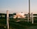 A shiny food trailer is parked on grassy terrain near a body of water during sunset. The trailer reflects the warm light, and there are signs and white posts surrounding the area. A wooden pole stands nearby, and distant trees line the horizon.