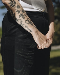 A close-up of a person's forearm with detailed black ink tattoos, featuring floral designs, scissors, and other objects. The person is wearing a white sleeveless top and dark pants.
