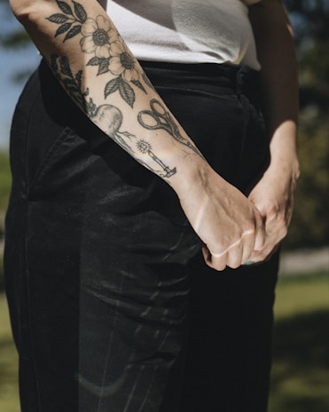 A close-up of a person's forearm with detailed black ink tattoos, featuring floral designs, scissors, and other objects. The person is wearing a white sleeveless top and dark pants.