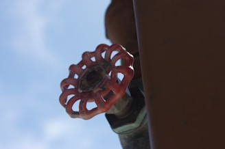 A close-up view of a red valve handle attached to a pipe against a backdrop of a clear blue sky. The metal valve is rusted and weathered, indicating it may be outdoors.