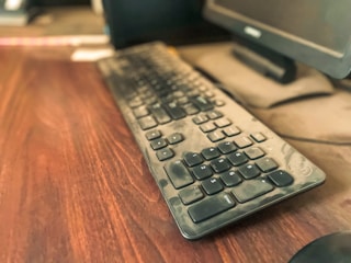 A dusty black keyboard is placed on a wooden surface next to a computer monitor. The dust on the keyboard is clearly visible, indicating that it hasn't been cleaned for a while. The background is blurred, focusing the attention on the keyboard.