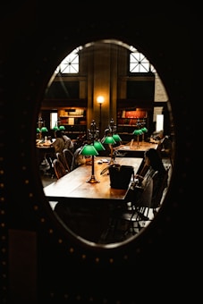 A cozy library room with dim lighting and a warm atmosphere. Wooden desks are adorned with classic green desk lamps. People are seated, quietly reading or working on laptops, surrounded by bookshelves filled with various books. The scene is framed by an oval window, adding a unique perspective.