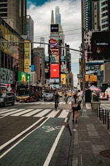 A bustling city street features tall buildings adorned with digital billboards and advertisements. People are walking along the sidewalks, and cyclists navigate the bike lane. In the background, numerous vehicles, including buses and taxis, move along the road.