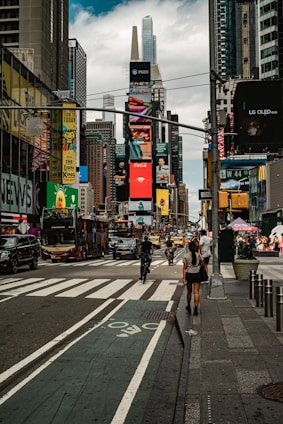 A bustling city street features tall buildings adorned with digital billboards and advertisements. People are walking along the sidewalks, and cyclists navigate the bike lane. In the background, numerous vehicles, including buses and taxis, move along the road.