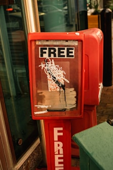 A red newsstand with a scratched and marked front glass displaying the word 'FREE' prominently at the top and bottom. It appears to be placed on a sidewalk near a building entrance.