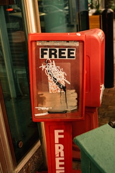 A red newsstand with a scratched and marked front glass displaying the word 'FREE' prominently at the top and bottom. It appears to be placed on a sidewalk near a building entrance.