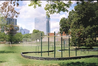 An urban park setting with a playground featuring climbing structures bordered by lush green grass. Tall modern skyscrapers in the background contrast with the natural environment. Trees provide some shade, and a clear blue sky with scattered clouds enhances the serene atmosphere.