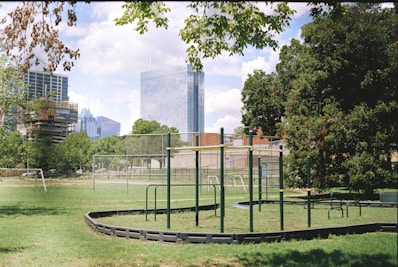 An urban park setting with a playground featuring climbing structures bordered by lush green grass. Tall modern skyscrapers in the background contrast with the natural environment. Trees provide some shade, and a clear blue sky with scattered clouds enhances the serene atmosphere.