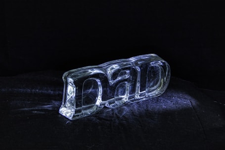 A clear glass sculpture spelling out the word 'DAD' is placed on a dark surface, illuminated by a focused light source creating reflections and shadows.