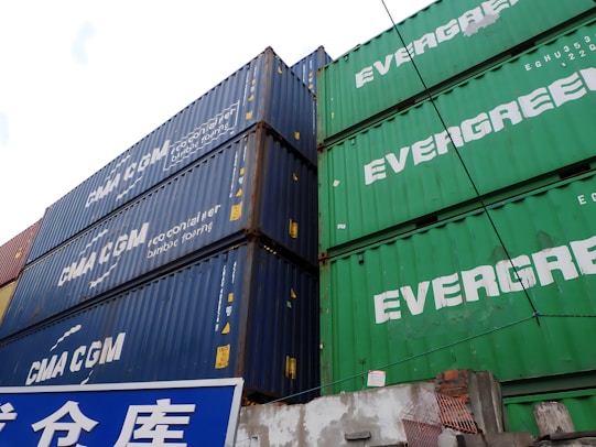 Several stacked shipping containers, predominantly showcasing the brands 'CMA CGM' on blue containers and 'EVERGREEN' on green containers. Below the containers is a blue and white sign with Asian characters.