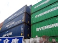 Several stacked shipping containers, predominantly showcasing the brands 'CMA CGM' on blue containers and 'EVERGREEN' on green containers. Below the containers is a blue and white sign with Asian characters.