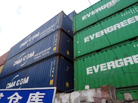 Several stacked shipping containers, predominantly showcasing the brands 'CMA CGM' on blue containers and 'EVERGREEN' on green containers. Below the containers is a blue and white sign with Asian characters.