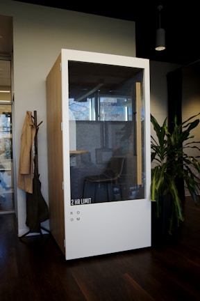 A modern, phone booth-style meeting pod is situated in an office environment. Made of wood with a large glass front, it has a label '2 HR LIMIT' and 'ROOM' on the lower section. Inside, a simple chair and a bit of office décor are visible. The booth is placed next to a coat rack with hanging items and a potted plant, all set on a dark wooden floor.