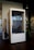 A modern, phone booth-style meeting pod is situated in an office environment. Made of wood with a large glass front, it has a label '2 HR LIMIT' and 'ROOM' on the lower section. Inside, a simple chair and a bit of office décor are visible. The booth is placed next to a coat rack with hanging items and a potted plant, all set on a dark wooden floor.