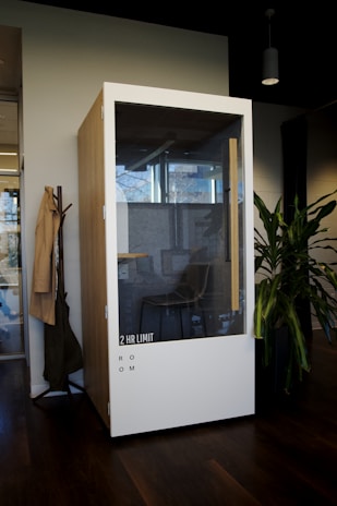 A modern, phone booth-style meeting pod is situated in an office environment. Made of wood with a large glass front, it has a label '2 HR LIMIT' and 'ROOM' on the lower section. Inside, a simple chair and a bit of office décor are visible. The booth is placed next to a coat rack with hanging items and a potted plant, all set on a dark wooden floor.