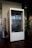 A modern, phone booth-style meeting pod is situated in an office environment. Made of wood with a large glass front, it has a label '2 HR LIMIT' and 'ROOM' on the lower section. Inside, a simple chair and a bit of office décor are visible. The booth is placed next to a coat rack with hanging items and a potted plant, all set on a dark wooden floor.