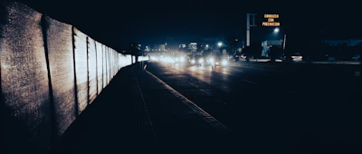 A nighttime scene featuring a dimly lit road with several vehicles approaching. Their headlights create a trail of light, illuminating the dark surroundings. On the right, a road sign with the text 'Conduzca con Precaución' is visible, indicating to drive with caution. The left side of the image has a barrier or wall reflecting the headlights’ glow.