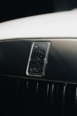 A close-up of a Rolls-Royce car emblem set against a shiny, metallic background. The emblem features the iconic double 'R' logo, indicative of luxury and elegance. The surface appears well-polished, enhancing the premium feel of the image.