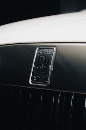 A close-up of a Rolls-Royce car emblem set against a shiny, metallic background. The emblem features the iconic double 'R' logo, indicative of luxury and elegance. The surface appears well-polished, enhancing the premium feel of the image.
