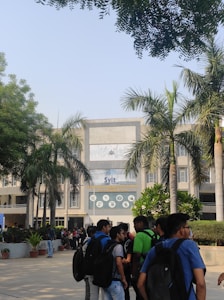 A group of students standing and chatting in front of a large academic building with a prominent entrance. The building is surrounded by palm trees, and the architecture is modern, featuring a large central display with the name 'Svit'. The sunny day casts clear shadows, and the atmosphere seems lively and bustling.