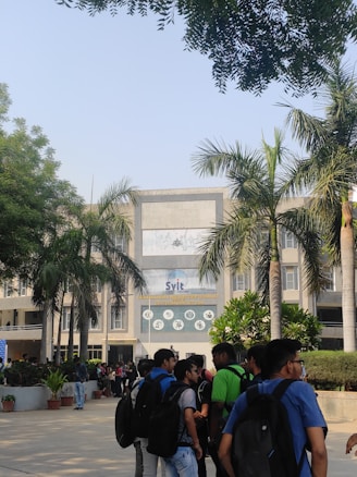 A group of students standing and chatting in front of a large academic building with a prominent entrance. The building is surrounded by palm trees, and the architecture is modern, featuring a large central display with the name 'Svit'. The sunny day casts clear shadows, and the atmosphere seems lively and bustling.