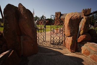 A rustic, ornate metal gate adorned with cross symbols stands open, leading into an area with a stone building resembling a castle. Large, reddish rock formations frame the entrance under a clear blue sky. Green grass and flags can be seen in the background.