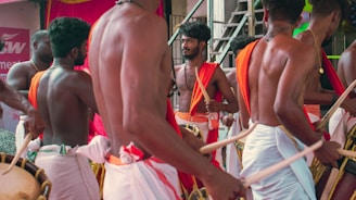 A group of men dressed in traditional attire play percussion instruments. They are gathered closely, some facing the camera, others with their backs turned. The clothing comprises mostly white garments with orange and red fabric draped over their shoulders. The setting appears festive, and the men seem focused on their performance.