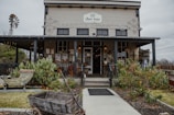 A rustic, two-story building with a wooden exterior and a small porch. The sign on top indicates it is a home furnishings store. The facade is adorned with string lights and decorative elements. In the foreground, there are various plants including cacti, an old wooden wheelbarrow, and a windmill to the side.