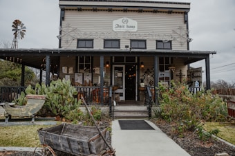 A rustic, two-story building with a wooden exterior and a small porch. The sign on top indicates it is a home furnishings store. The facade is adorned with string lights and decorative elements. In the foreground, there are various plants including cacti, an old wooden wheelbarrow, and a windmill to the side.