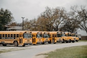 A group of yellow school buses parked in a row on a gravel path next to grassy areas. The buses have a logo reading 'Rockin' R River Rides' displayed across their sides. In the background, there are bare trees and a building partially visible.