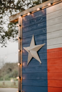 A wooden wall painted with the Texas flag design featuring vertical stripes of blue, white, and red, with a prominent white star in the middle of the blue section. The edge of the structure is adorned with small circular string lights. Trees and foliage are faintly visible in the blurred background, contributing to an outdoor setting.