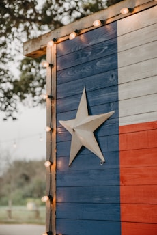 A wooden wall painted with the Texas flag design featuring vertical stripes of blue, white, and red, with a prominent white star in the middle of the blue section. The edge of the structure is adorned with small circular string lights. Trees and foliage are faintly visible in the blurred background, contributing to an outdoor setting.