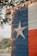 A wooden wall painted with the Texas flag design featuring vertical stripes of blue, white, and red, with a prominent white star in the middle of the blue section. The edge of the structure is adorned with small circular string lights. Trees and foliage are faintly visible in the blurred background, contributing to an outdoor setting.