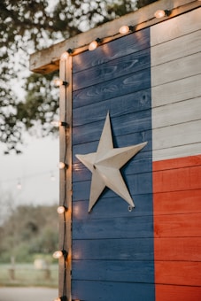A wooden wall painted with the Texas flag design featuring vertical stripes of blue, white, and red, with a prominent white star in the middle of the blue section. The edge of the structure is adorned with small circular string lights. Trees and foliage are faintly visible in the blurred background, contributing to an outdoor setting.